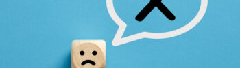 Unhappy face icon on a wooden cube with cross x sign in a speech bubble.